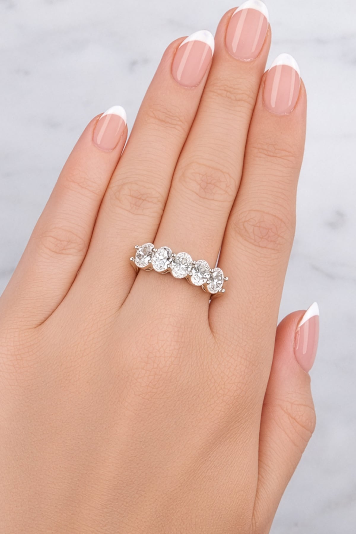 Silver ring with five diamonds on a hand with light pink nail polish against a light grey background at LeGassick.