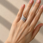 A close-up of a hand wearing a delicate white gold ring set with small clear diamonds, photographed against a warm beige background with soft diagonal shadows.