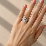 A close-up of a hand wearing a delicate white gold ring set with small clear diamonds, photographed against a warm beige background with soft diagonal shadows.