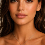 A close-up portrait of a woman wearing a delicate white gold cross pendant necklace with green emerald gemstones and diamonds, set against a soft neutral background.
