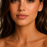 A close-up portrait of a woman wearing a delicate white gold cross pendant necklace with green emerald gemstones and diamonds, set against a soft neutral background.