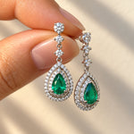 Fingers holding a pair of 18ct white gold Colombian emerald and diamond drop earrings featuring pear-shaped green emeralds surrounded by double halos of round brilliant-cut diamonds.