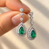 Fingers holding a pair of 18ct white gold Colombian emerald and diamond drop earrings featuring pear-shaped green emeralds surrounded by double halos of round brilliant-cut diamonds.