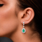 A woman wearing a pair of 18ct white gold Colombian emerald and diamond drop earrings featuring pear-shaped green emeralds surrounded by double halos of round brilliant-cut diamonds.