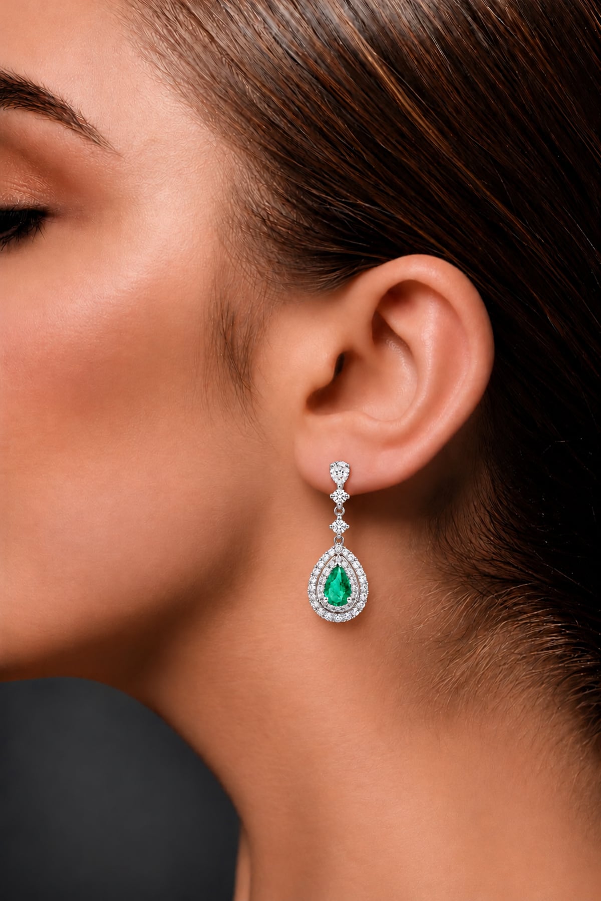 A woman wearing a pair of 18ct white gold Colombian emerald and diamond drop earrings featuring pear-shaped green emeralds surrounded by double halos of round brilliant-cut diamonds.