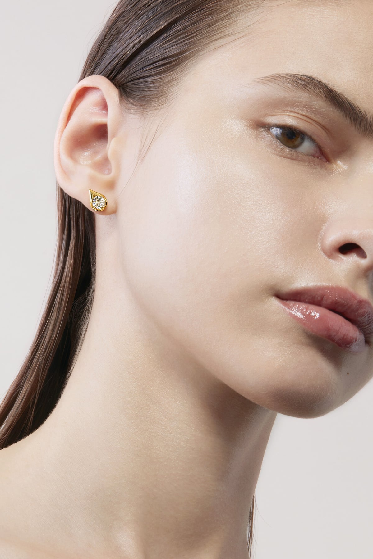 Close-up of a woman wearing a gold earring with a diamond on a neutral background