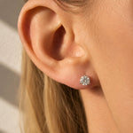 A close-up of a woman's ear wearing a round brilliant-cut diamond stud earring. The image is shot in warm, direct sunlight with soft shadows on a light background.