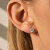 A close-up of a woman's ear wearing a round brilliant-cut diamond stud earring. The image is shot in warm, direct sunlight with soft shadows on a light background.