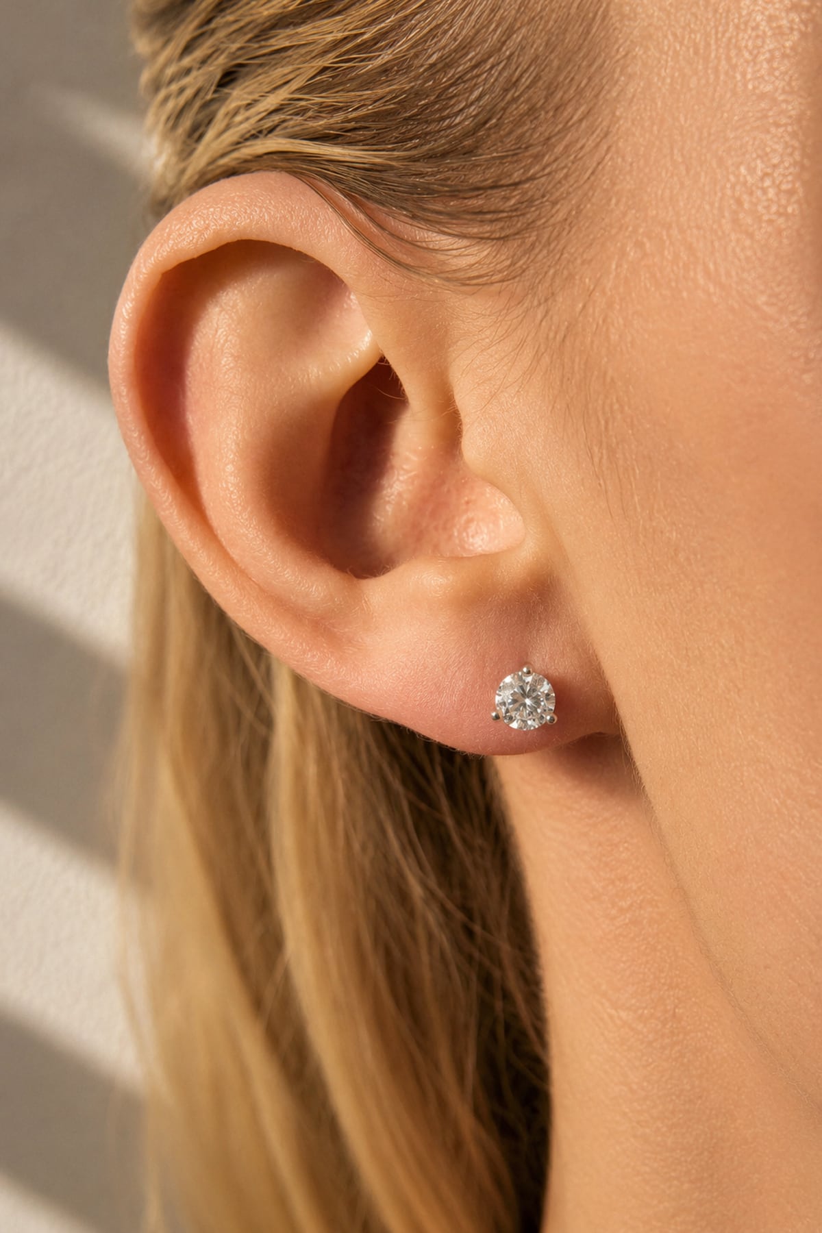 A close-up of a woman's ear wearing a round brilliant-cut diamond stud earring. The image is shot in warm, direct sunlight with soft shadows on a light background.