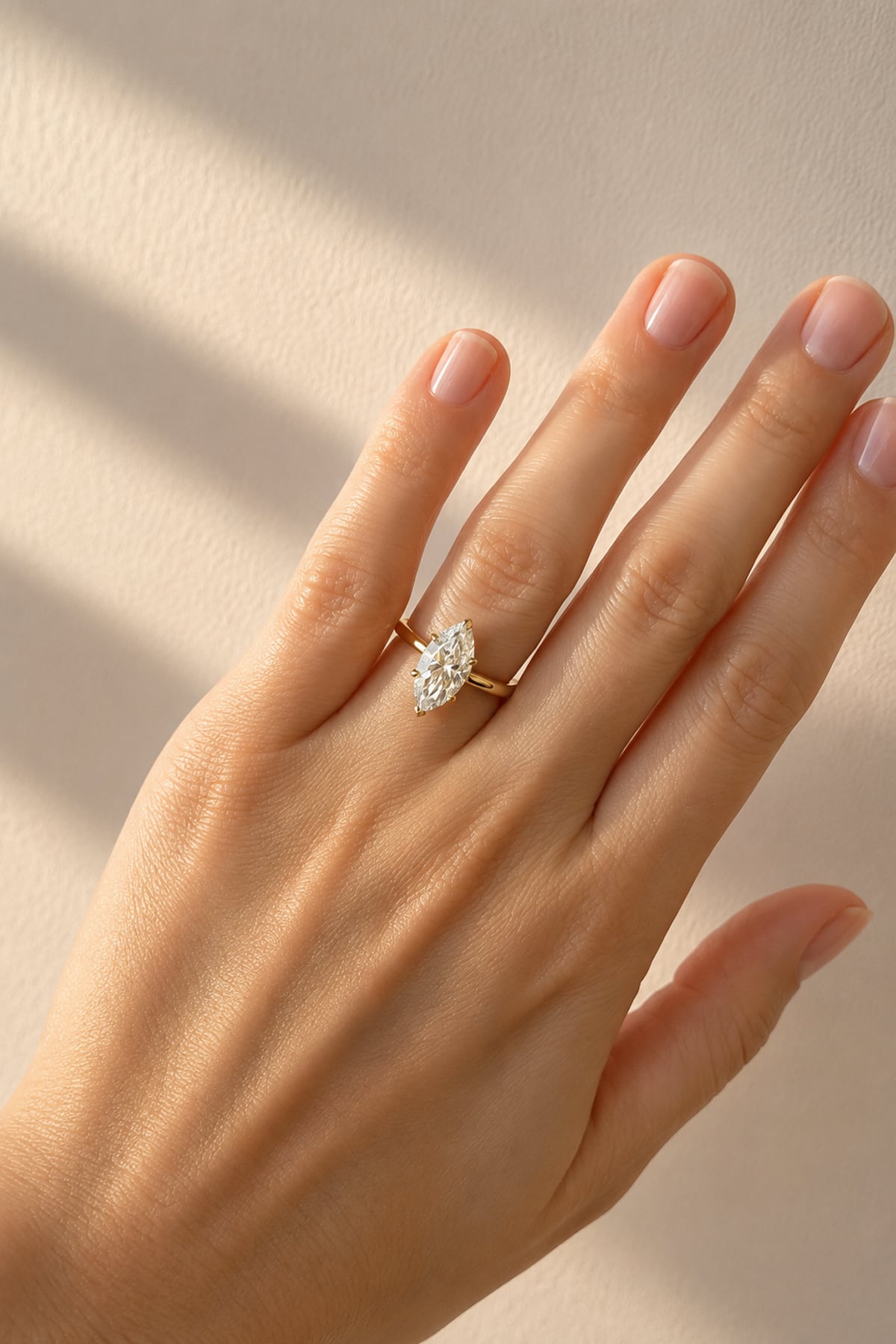 A close-up of a hand wearing a gold ring with a marquise-cut clear lab diamond, set against a warm beige background with soft window-light shadows.