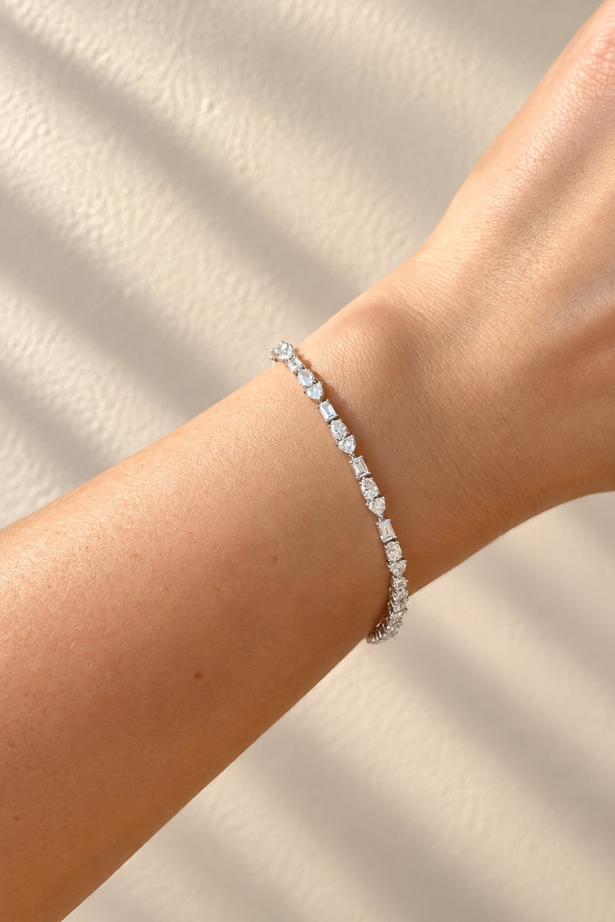 A delicate platinum tennis bracelet with various shaped lab dimaonds, worn on a person's wrist against a textured beige background with soft shadow overlays.
