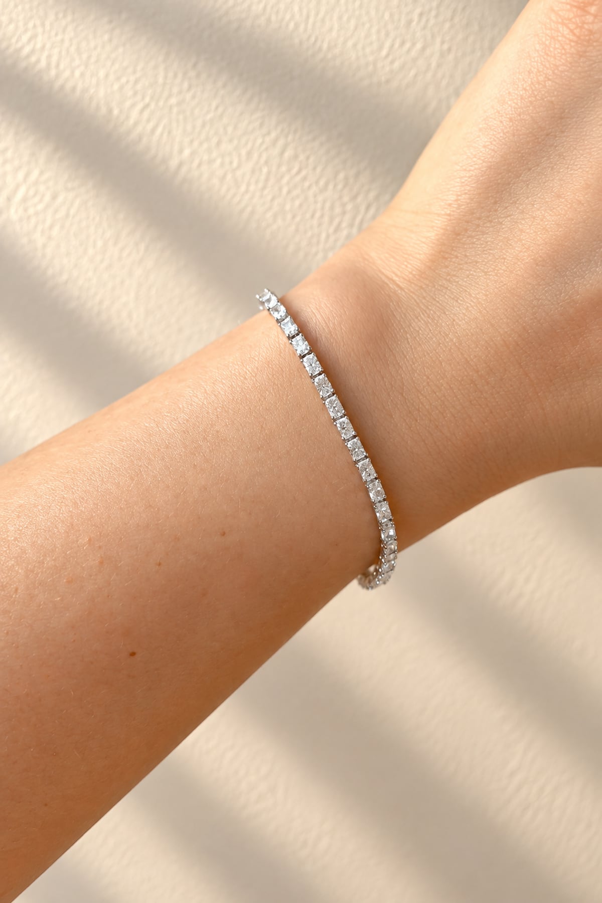 A slim tennis bracelet with clear rectangular diamonds worn on a person's wrist, shown against a warm beige textured background with soft shadow overlays.