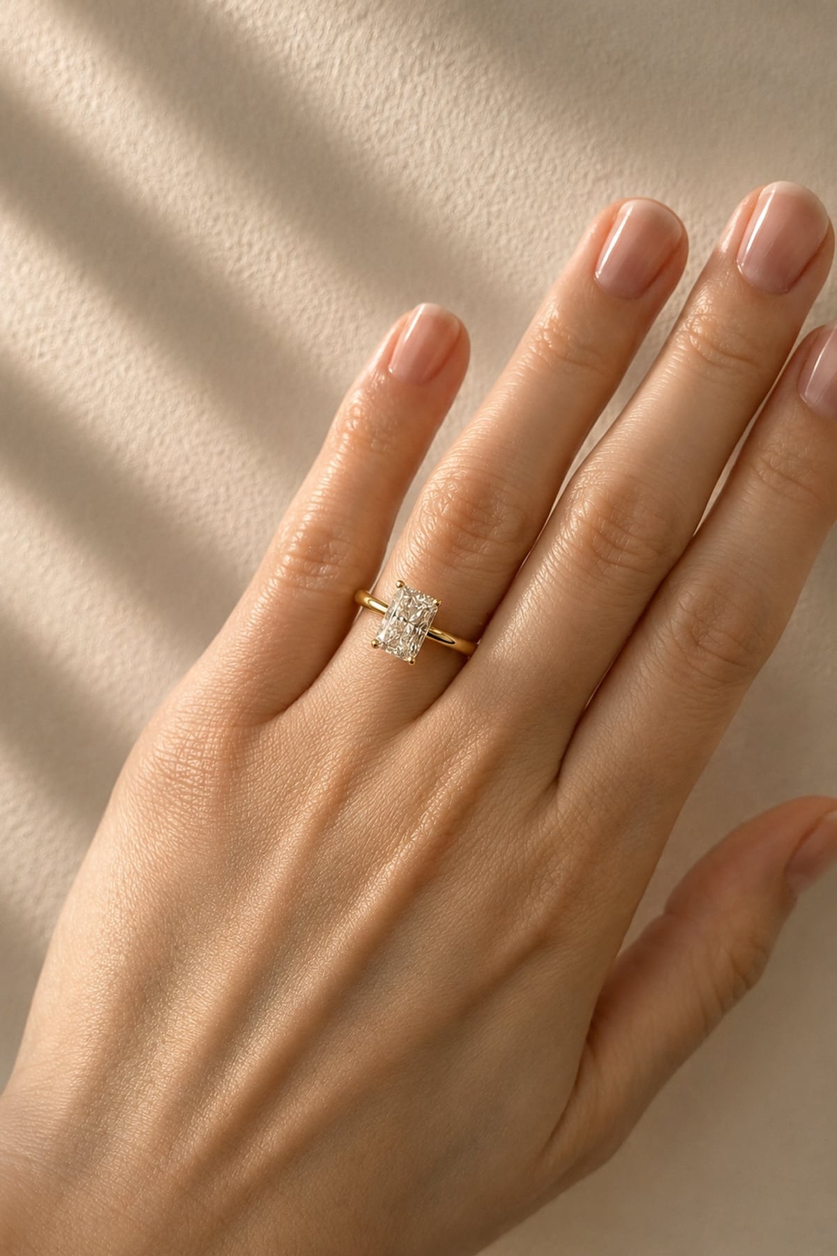 A close-up of a hand wearing a yellow gold ring with a rectangular clear lab diamond, set against a warm beige background with soft diagonal shadows.